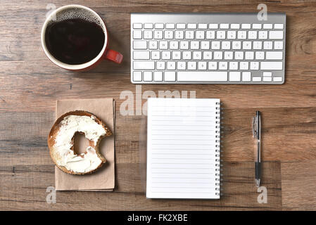 Ein Bagel mit Frischkäse und einen Imbiss neben einer Computer-Tastatur und einen Block und Stift herausgenommen. Stockfoto