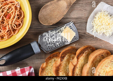 Draufsicht auf einen Küchentisch mit einem Teller Spaghetti, Knoblauchbrot, geriebenen Parmesan, Reibeisen, hölzerne Löffel, eine rote ch Stockfoto