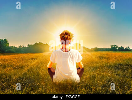 Rückansicht des junge blonde Frau im Feld meditieren vor der Sonne zu sitzen. Stockfoto