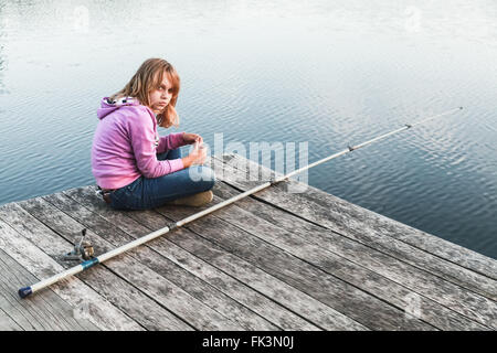 Blonde kaukasische Mädchen sitzen auf einem hölzernen Pier mit Angelrute Stockfoto