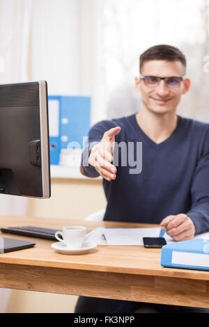 Lächelnde Jungunternehmer in Gläsern streckt seine Hand für einen Händedruck an seinem Arbeitsplatz Stockfoto