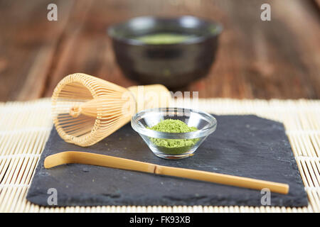Grünen Matcha-Pulver, Tee und Schneebesen und Löffel für die Vorbereitung auf schwarzen Steinplatte und weißen Tisch Bambusmatte Stockfoto