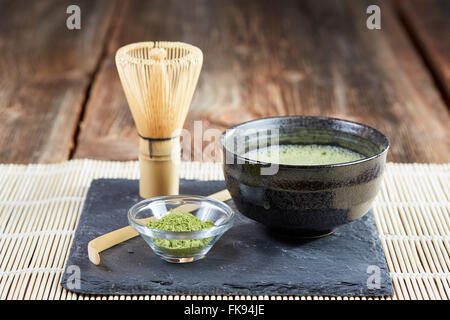 Grünen Matcha-Pulver, Tee und Schneebesen und Löffel für die Vorbereitung auf schwarzen Steinplatte und weißen Tisch Bambusmatte Stockfoto