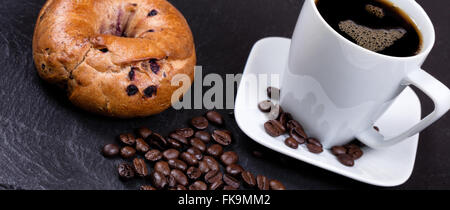 Seitlicher Blick auf gerösteten Kaffeebohnen auf schwarzem Schiefer Naturstein mit Kaffee trinken und Bagel im Hintergrund. Stockfoto