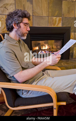 Mittleren Alters kaukasischen männlichen sitzen in der Nähe einen Kamin, einen akademischen oder finanziellen Bericht lesen zu unterscheiden. Stockfoto