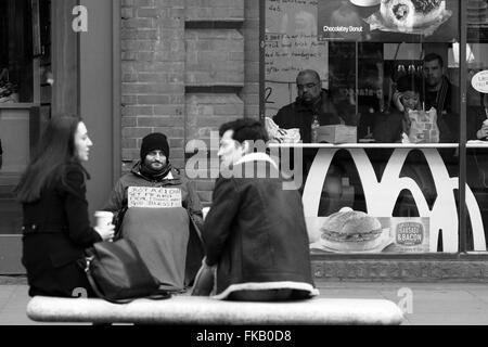 Zwei Menschen sitzen und trinken, während ein Obdachloser Mann neben einem Mcdonalds-Restaurant in Oxford Street, London, England sitzt Stockfoto