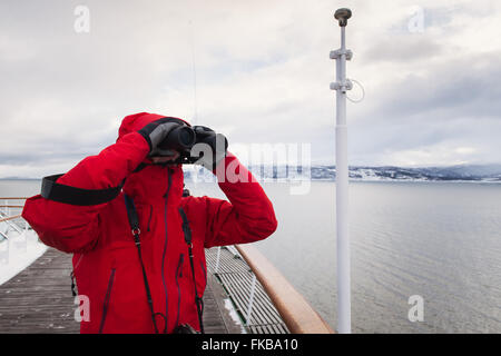 Ein Mann in einem roten Mantel verwendet Fernglas auf dem Deck eines Kreuzfahrtschiffes in Arktis Norwegen. Stockfoto