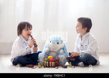 Zwei entzückende kleine Kinder, junge Brüder, mit Spaß Essen Schokohasen und mit Eiern zu Hause spielen, Hintergrundbeleuchtung Stockfoto