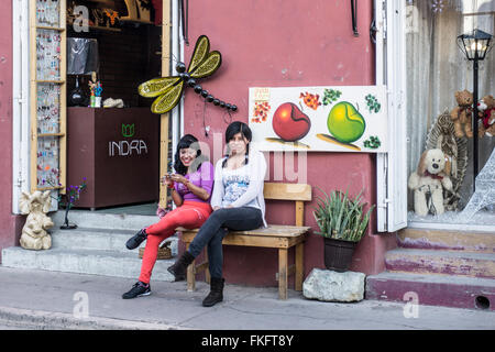 2 freche junge mexikanische Shop Begleiter Mädchen draußen auf Bürgersteig Bank vor Neuheit Boutique wo arbeiten sie Oaxaca Stockfoto