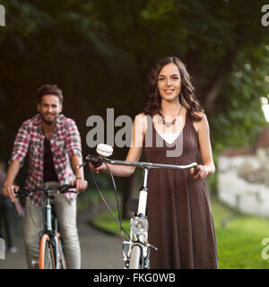 schöne junge Frau neben das Motorrad in den Vordergrund und Hintergrund gut aussehend männlichen mit dem Fahrrad auf einer Gasse mit grünen Bäumen Stockfoto