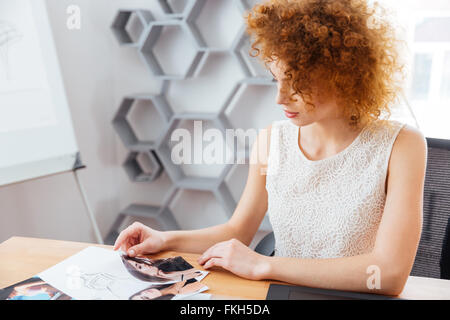 Ziemlich lockig rothaarige junge Frau Modedesigner sitzen und wählen Sie Modelle auf Fotos im Büro Stockfoto