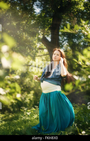 Schöne schwangere Frau im blauen Kleid in grün Sommerpark Natur und Sonne genießen. Voller Höhe Portrait, Blick in die Kamera. Stockfoto