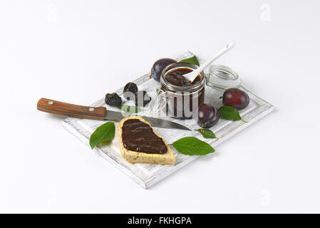 Glas Pflaumenmus, frische und getrocknete Pflaumen und Scheibe Baguette auf Holzbrett Stockfoto