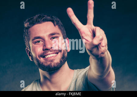 Porträt von glücklich lächelnde gut aussehender Mann Kerl geben Frieden Sieg V Zeichen Geste im Studio auf schwarz. Stockfoto