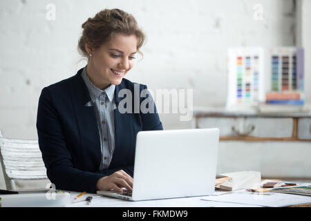 Porträt von schönen glücklich lächelnden jungen Designer Frau auf Laptop am Schreibtisch arbeiten. Attraktives fröhlich Modell Stockfoto