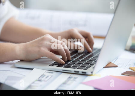 Arme von schönen jungen Designer Frau Tippen auf Laptop Schreibtisch zu Hause sitzen bedeckt in Zeichnungen und Baupläne Stockfoto