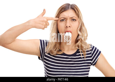Blonde Frau mit einem Bündel von Zigaretten in den Mund mit einer Handpistole auf den Kopf, die isoliert auf weißem Hintergrund Stockfoto