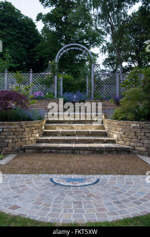 Mosaik-Kunst, Pflasterarbeiten, Blütenpflanzen, Schritte und Rankgitter Bogen in der Dämmerung - gestaltete, gepflegten Garten, Burley in Wharfedale, Yorkshire, England. Stockfoto