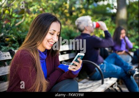 Junge Frau sitzt auf der Parkbank Smartphone Texte lesen Stockfoto