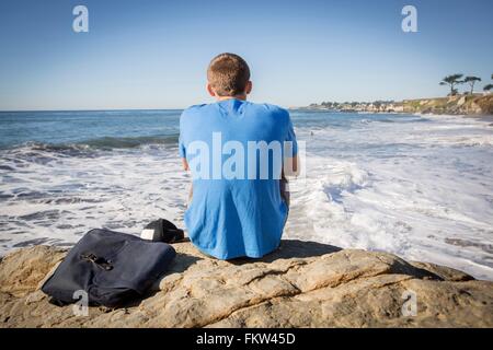 Junger Mann sitzen von Felsen, Blick auf das Meer, Rückansicht Stockfoto