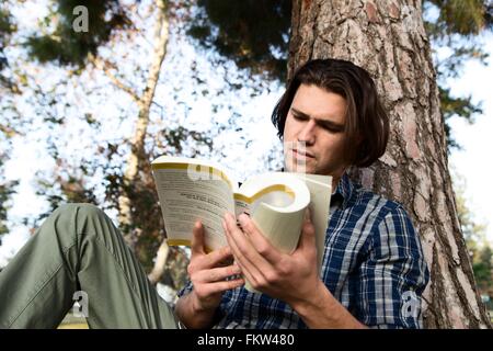 Niedrigen Winkel Ansicht der junge Mann sitzt gegen Baum Buch Stockfoto