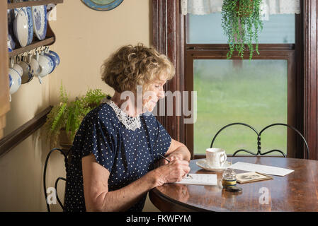 In einer historischen Szene aus dem 30er oder 40er Jahren schreibt eine attraktive ältere Frau einen Brief, als sie an einem Tisch am Fenster sitzt. Stockfoto