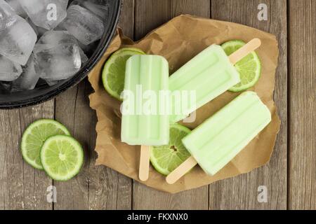 Hausgemachter Limetten Joghurt Eis am Stiel mit frischen Limetten auf Papier mit rustikalen Holz Hintergrund Stockfoto