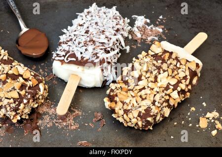 Hausgemachte Schokolade getaucht Eis am Stiel mit Nüssen und Kokos vor einem rustikalen dunklen Hintergrund. Stockfoto