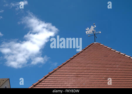 Wetterfahne mit Teufel und Indikator für die Himmelsrichtungen auf ein Spitzdach mit roten Dachziegeln vor blauem Himmel. Stockfoto