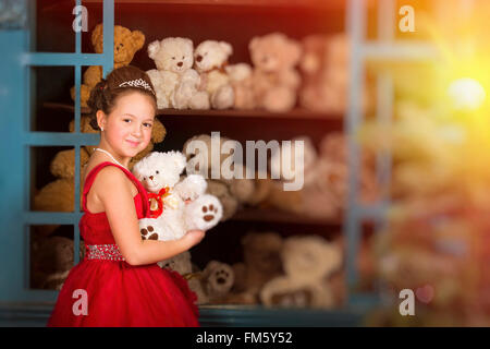 Entzückende kleine Mädchen, gekleidet in einem üppigen roten Kleid umarmt einen Teddybären neben dem Fenster mit Spielzeug Stockfoto