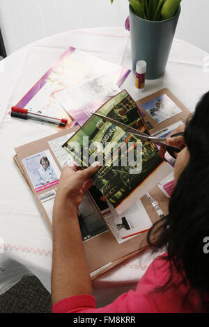 Hände halten bedrucktes Papier in der Hand und Schere in die andere Karte mit Artikeln und Bildern auf stecken, bleiben Stifte und Kleber im Hintergrund Stockfoto