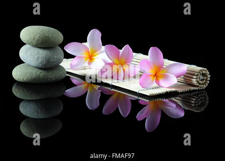 Wellness-, Beauty- und Wellness-Konzept - gestapelt Zen Steinen Frangipani-Blüten auf einer Bambusmatte mit Reflexion, dunklen Hintergrund. Stockfoto