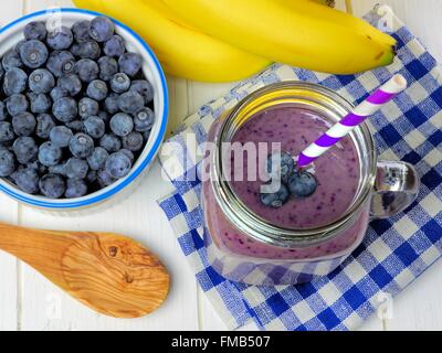 Heidelbeer-Smoothie über Ansicht mit Bananen, karierte Tuch und Löffel auf weißem Holz Stockfoto