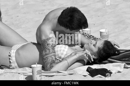 Reportageaufnahme attraktives Paar, Mann mit Tattoos, Frau im Bikini, Tassen trinken, liegen am Strand, Zuneigung, Liebe Stockfoto