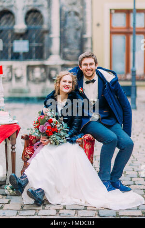 Fröhliche Bräutigam und die Braut sitzt auf Vintage Sessel glücklich lächelnd und Blick in die Kamera Stockfoto