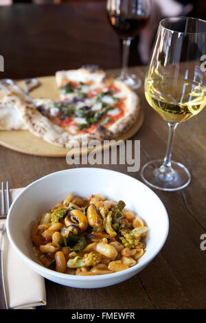 Eine Schale mit weißen Bohnen, Romanesco und Muscheln-Suppe mit Weißwein und eine Pizza auf dem Tisch hinter sich. Stockfoto