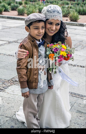 glückliche mexikanische Braut hält Frühlingsblumen & kniet um kleinen Neffen für Hochzeitsbild auf der Kirche Santo Domingo Plaza umarmen Stockfoto