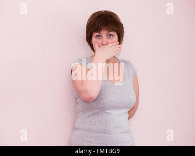 Portrait attractive mature woman with shocked, surprised, anxious facial expression, covering mouth with hand on plain backgroun Stockfoto