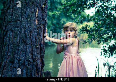 kleines blondes Mädchen mit langen Haaren lehnte sich an den Baumstamm Stockfoto