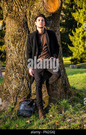 Hübscher junger Mann lehnte sich gegen Baum, Blick in die Kamera, an einem sonnigen Tag trägt einen schwarzen Mantel Stockfoto