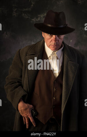 Unheimlicher aussehender Mann mit Mantel und Hut mit einer Hand über Gürtel Pistole Porträt Hintergrund in niedrigen zentrale Beleuchtung Stockfoto
