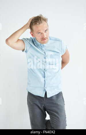 Brutaler Mann mit einem kleinen Bart lehnt an der Wand, trägt ein blaues Hemd mit kurzen Ärmeln und graue Hose, Hände sind hinter seinem Stockfoto