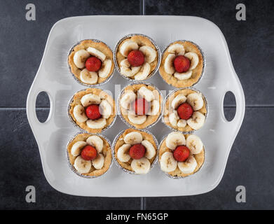 Neun Törtchen mit Erdbeeren und Bananen Scheiben auf weiße Schale und dunklen strukturierten Hintergrund. Ansicht von oben. Stockfoto