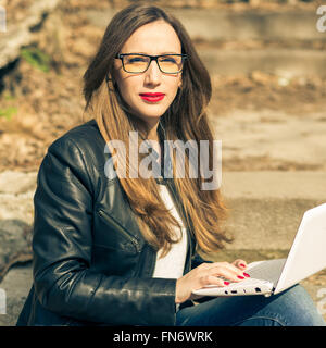 Junge, lächelnde Frau mit langen Haaren, die mit Laptop auf Treppe im Park. Warme Farbe getönt Bild Stockfoto