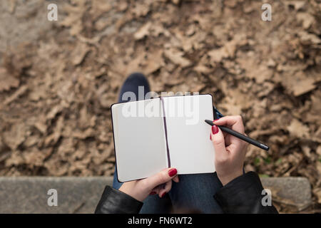 Ansicht von oben Bild des offenen Pad in Feamle Hände mit Stift auf Treppen im Park sitzen. Leere Blätter kleine Notebook-Draufsicht Stockfoto