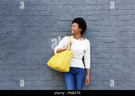 Porträt von lächelnden jungen Afrikanerin tragen einer Tasche stehen gegen eine graue Wand Stockfoto