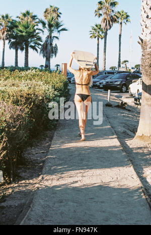 Eine Frau in einem schwarzen Bikini zu Fuß auf einem Weg ein Surfbrett auf dem Kopf tragen. Stockfoto