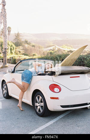 Eine junge Frau in Jeans-Shorts in ein Cabrio mit einem Surfbrett auf dem Rücksitz gelehnt. Stockfoto