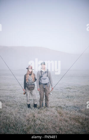 Zwei Menschen, die zu Fuß über eine Wiese im frühen Morgennebel tragen Angelruten. Stockfoto