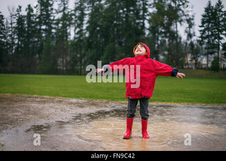 Kleiner Junge mit ausgestreckten Armen in Pfütze im Regen stehen Stockfoto